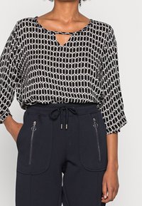 Blouse à motifs noir et blanc avec un col en trou de serrure, associée à un pantalon de survêtement bleu marine avec cordon de serrage et poches zippées.