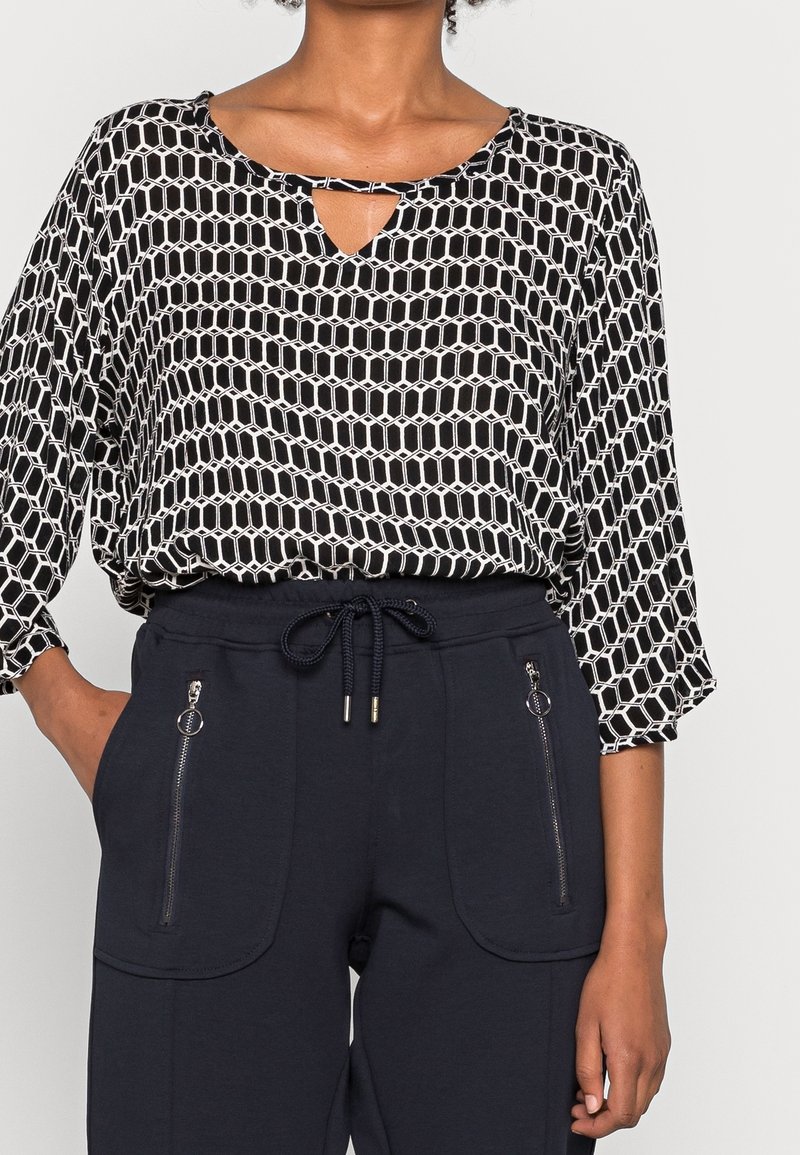 Blouse à motifs noir et blanc avec un col en trou de serrure, associée à un pantalon de survêtement bleu marine avec cordon de serrage et poches zippées.