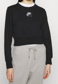 Osoba oblečená v černé zkrácené mikině Nike Air s bílým tričkem a světle šedými tepláky na stahování, viditelné torzo a ruce.