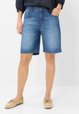 Person trägt blaue, knielange Jeansshorts, schwarzes Top, beige Schnür-Loafer und ein silbernes Armband, steht mit einer Hand in der Tasche.