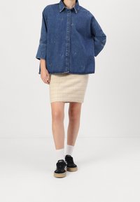 Persona che indossa una camicia di denim blu oversize, una gonna beige a lunghezza ginocchio con trama, calzini bianchi e sneakers nere su uno sfondo bianco semplice.