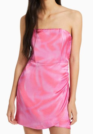 Frau trägt ein trägerloses glänzendes pinkfarbenes Minikleid mit gerafftem Detail am Rock, steht mit locker an den Seiten hängenden Armen.