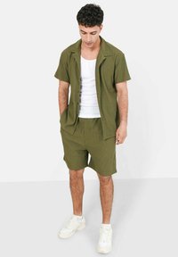 Donkergroen shirt met korte mouwen en bijpassende shorts, gemaakt van textuurstof. Witte tanktop eronder, gecombineerd met witte sportschoenen.