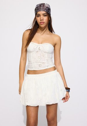 Młoda kobieta ubrana w biały top bez ramiączek z ażurowym wzorem i dopasowaną spódnicę, z czarno-białą, wzorzystą bandaną na głowie i bransoletkami.