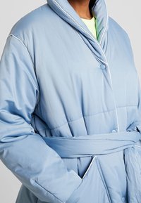 Accappatoio imbottito azzurro chiaro con colletto a scialle, presenta dettagli in bianco, una cintura da annodare e chiusure a bottoni, progettato per comfort e calore.