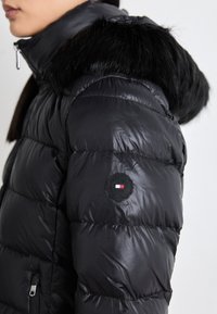 Giacca a vento nera con una texture lucida e trapuntata. Presenta un bordo in pelliccia sintetica sul cappuccio e una piccola patch con logo sulla manica.