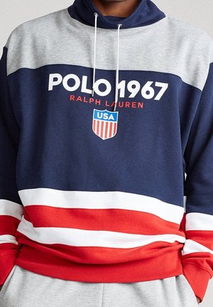 Granatowo-niebieska, szara, czerwona i biała bluza z kapturem z tekstem "POLO 1967 RALPH LAUREN" oraz emblematem tarczy USA na piersi, noszona przez osobę.