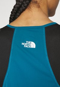 Camiseta de deporte con tejido de malla en teal en la parte trasera, detalles en negro y un logo blanco en la esquina superior izquierda. Costuras planas y cuello redondo.