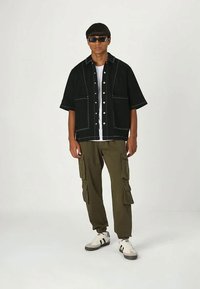 Black short-sleeve button-up shirt with white stitching, olive cargo pants, and white sneakers with black accents worn by a model.