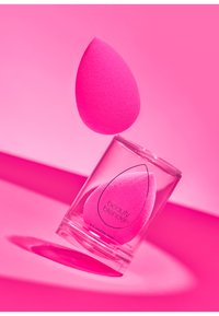 Spugna da trucco rosa a forma di lacrima, esposta in posizione verticale in un contenitore cilindrico trasparente con un marchio nero sul lato.