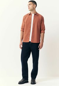 Chemise boutonnée couleur rouille sur un t-shirt blanc, associée à un pantalon bleu marine et des bottes noires. Tissu lisse, coupe décontractée et poche visible.