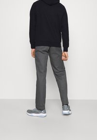 Uomo in piedi con le spalle alla fotocamera, che indossa un hoodie nero, pantaloni grigi e sneaker grigie con suola bianca, su uno sfondo chiaro e neutro.