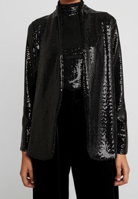 Černá pailletová bunda s uvolněným střihem, dlouhými rukávy a otevřeným předním designem. Hladká textura s třpytivým povrchem po celém obvodu.