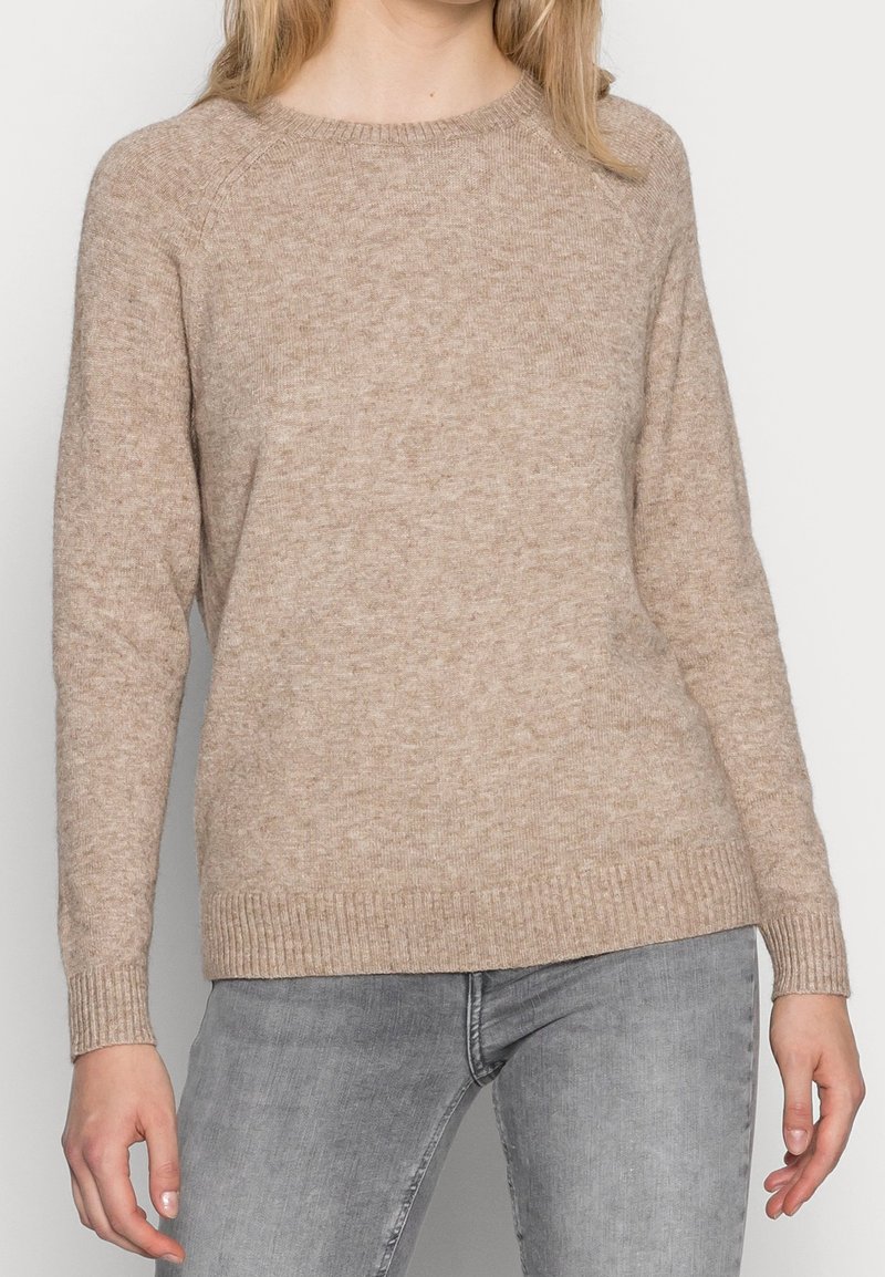 Femme portant un pull en tricot beige à manches longues avec des poignets côtelés, associé à un jean gris clair, debout devant un fond uni.