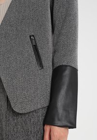 Šedý látkový blazer s vzorem houndstooth, který má černý kožený manžetový rukáv a zapínací kapsu na přední straně.