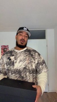 Schwarze rechteckige Box, die in der Hand gehalten wird, mit einem strukturierten, gemusterten Grafik-T-Shirt in hellen und dunklen Farbtönen und einem langärmeligen Unterhemd.