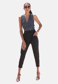 Zwarte, aansluitende broeken gecombineerd met een mouwloos, navy blouse met stippen, voorzien van een diepe V-hals en een wikkelontwerp. Accessoires omvatten een zonnebril.