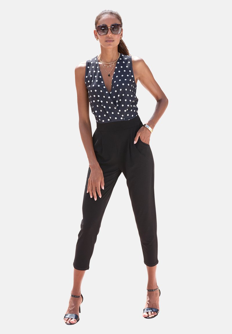 Zwarte, aansluitende broeken gecombineerd met een mouwloos, navy blouse met stippen, voorzien van een diepe V-hals en een wikkelontwerp. Accessoires omvatten een zonnebril.