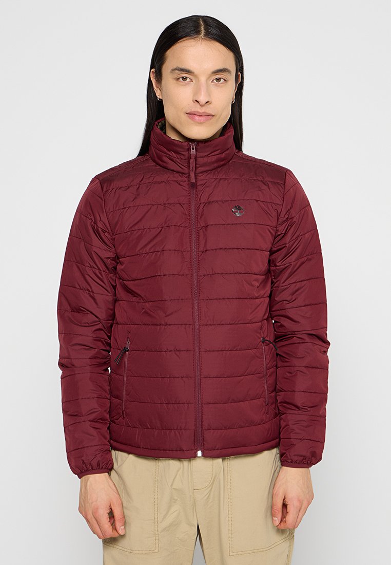 Giubbotto imbottito bordeaux con colletto alto, cuciture orizzontali, due tasche laterali con zip e un piccolo logo sul petto.