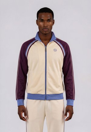 Homme portant une veste de survêtement crème et violette zippée avec des finitions bleues et un pantalon crème assorti sur un fond clair uni.