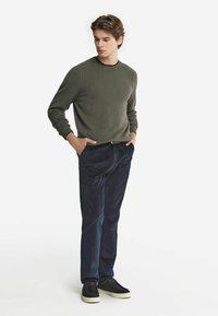 Grön stickad tröja med ribbade muddar, i kombination med marinblå kordbyxor. Svarta skor med vit sula. Modellen står med händerna i fickorna.