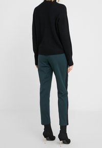 Pull noir côtelé associé à un pantalon bleu sarcelle avec des bandes latérales noires. Le pantalon est ajusté, et le modèle porte des bottines noires à talons.