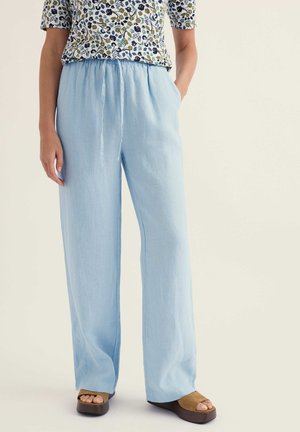 Személy világoskék, laza nadrágot visel, amelynek rugalmas derékpántja van, valamint virágmintás rövid ujjú felsőt, kezét zsebében tartja, barnás platform szandál van a lábán.