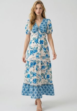Mujer joven caminando con vestido blanco hasta los tobillos con diseño azul floral y estampado, y mangas cortas abullonadas.
