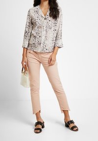 Kvinna klädd i en blus med ormtryck, ljusrosa cropped jeans med fransar, svarta klacksandaler och bär en genomskinlig handväska med trähandtag.