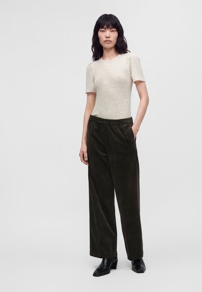 Femme aux cheveux noirs portant un haut beige à manches courtes, un pantalon large foncé et des bottines noires à talons, debout avec une main dans la poche.