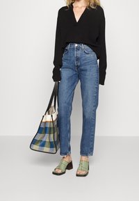 Schwarzer V-Ausschnitt-Pullover, hochtaillierte blaue Jeans, grüne Blockabsatzsandalen und eine karierte blau-grüne Tragetasche.