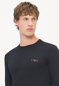 Black long-sleeve shirt in soft material featuring a small red logo on the chest, round neck design, and fitted silhouette.
