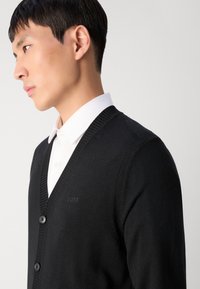 Cárdigan negro con escote en V, dos botones y detalles acanalados. Usado sobre una camisa blanca con cuello, que presenta un discreto bordado del logo en el pecho.