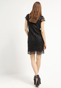 Kvinna med långt hår som bär en svart spetsklänning med kort ärm och svarta högklackade skor, stående med ryggen mot kameran.