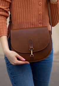 Brūna ādas crossbody soma ar noapaļotu formu, zelta metāla sprādzi un kontrastējošu šuvumu. Valkāta uz džinsu tērptas rokas.