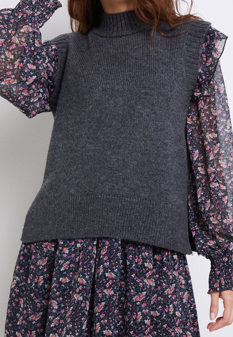 Femme portant un gilet en maille sans manches gris foncé sur une robe noire à fleurs à manches longues avec des poignets et des épaules à volants.