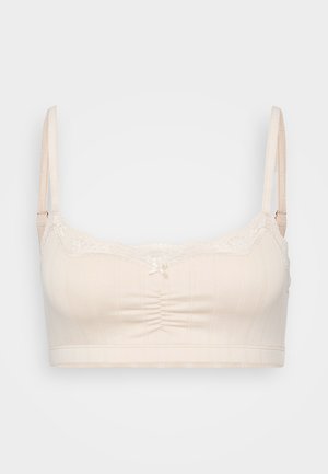 Vaaleanbeige bralette, jossa on pitsireunus, röyhelö keskiosassa ja säädettävät hihnat. Sisältää ribbikuvion ja pienen nauhatyylisen koristeen edessä.