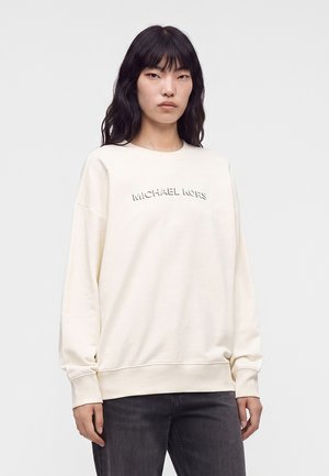 Donna con lunghi capelli neri, indossa una felpa bianca Michael Kors e jeans neri, in piedi su uno sfondo chiaro e semplice.