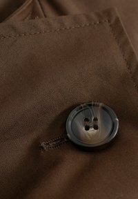 Detailný záber na tmavohnedé gombík prišitý na zodpovedajúcu hnedú látku so viditeľným stehom a štruktúrou.