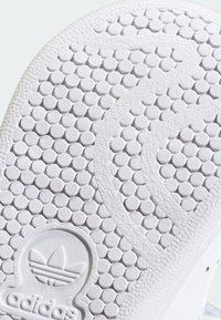 Biała gumowa podeszwa buta z okrągłym wzorem bieżnika i wytłoczonym logo Adidas w pobliżu pięty.