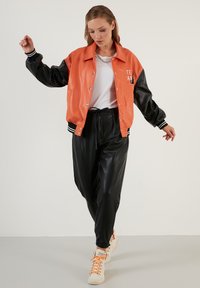 Oranje en zwarte neplederen jas met gestreepte manchetten, letterdetail op de borst, gecombineerd met zwarte neplederen broek en beige sneakers.