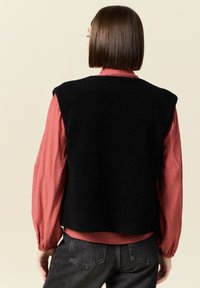 Svart fleeceväst med en kortad design, med breda axelöverdrag, lagd över en ribbad långärmad tröja i rosa, tillsammans med mörka jeans.