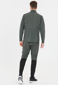 Grön idrottsjacka med en vadderad rygg, i kombination med tapered byxor med svarta detaljer. Materialet verkar lätt och elastiskt.