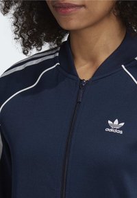 Osoba v námořnickém modrém zipovém svetru Adidas s bílými pruhy na ramenou a logem Adidas na hrudi.