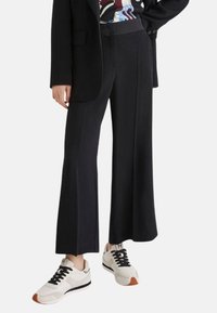 Pantaloni neri a gamba larga con una texture liscia, abbinati a sneaker crema con lacci neri e un accento marrone sulla suola.