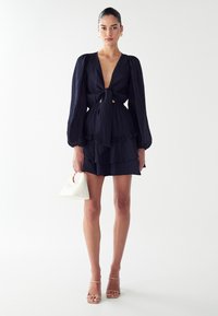 Mornarska obleka z dolgimi pufastimi rokavi, globokim V-izrezom, sprednjim trakom za zavezovanje in volančki na robu. Model drži belo geometrijsko torbico in nosi nude strap-on petke.