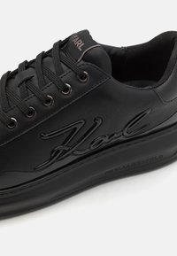 Zwarte sneaker met een gladde, gestructureerde bovenkant, vetersluiting, reliëflogo en een dikke rubberen zool. Minimalistische stijl met metalen accenten.
