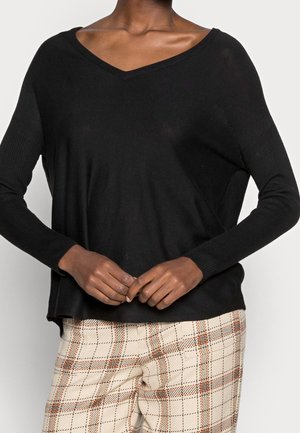 Personne portant un pull noir à col en V et manches longues, avec un pantalon à carreaux beige, debout les mains jointes à la taille.