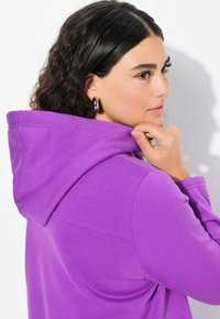 Sudadera con capucha morada de textura de tela suave, con cordón y bolsillo tipo canguro. La modelo está ajustando la capucha con la mano.