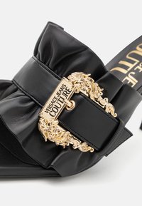 Zwarte leren slidesandaal met een strikdetail en een goudkleurige decoratieve gesp met "Versace Jeans Couture" branding.